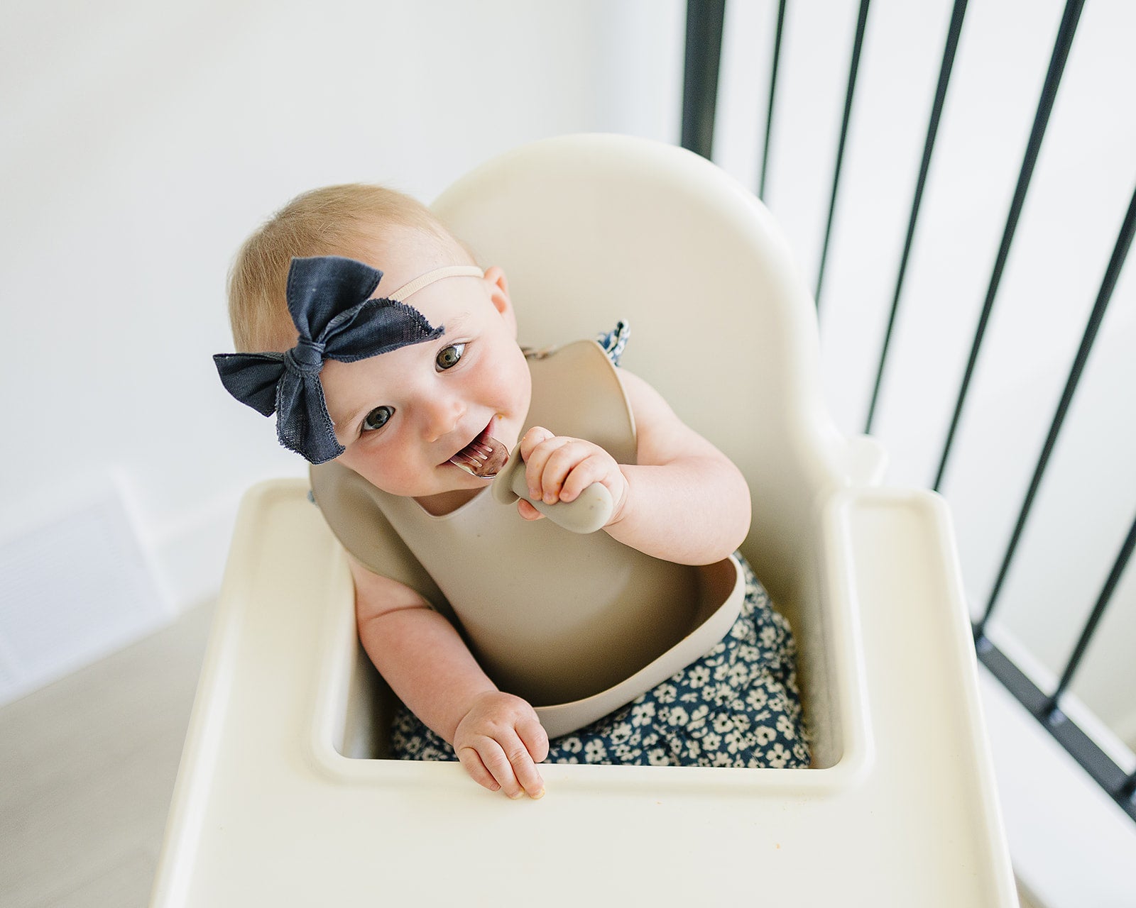 Baby sitting in a high chair wearing a silicon bib and floral pants with a navy bow headband.