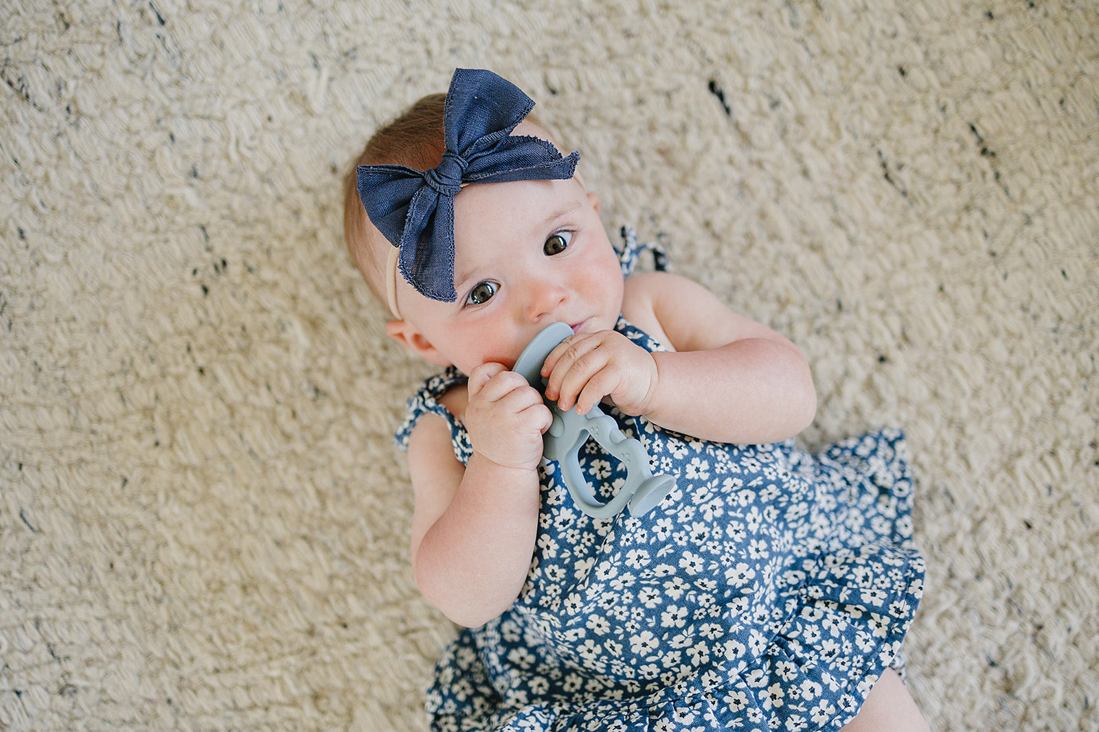 Baby lying on a carpeted floor wearing a blue floral dress and headband, holding a teething ring.