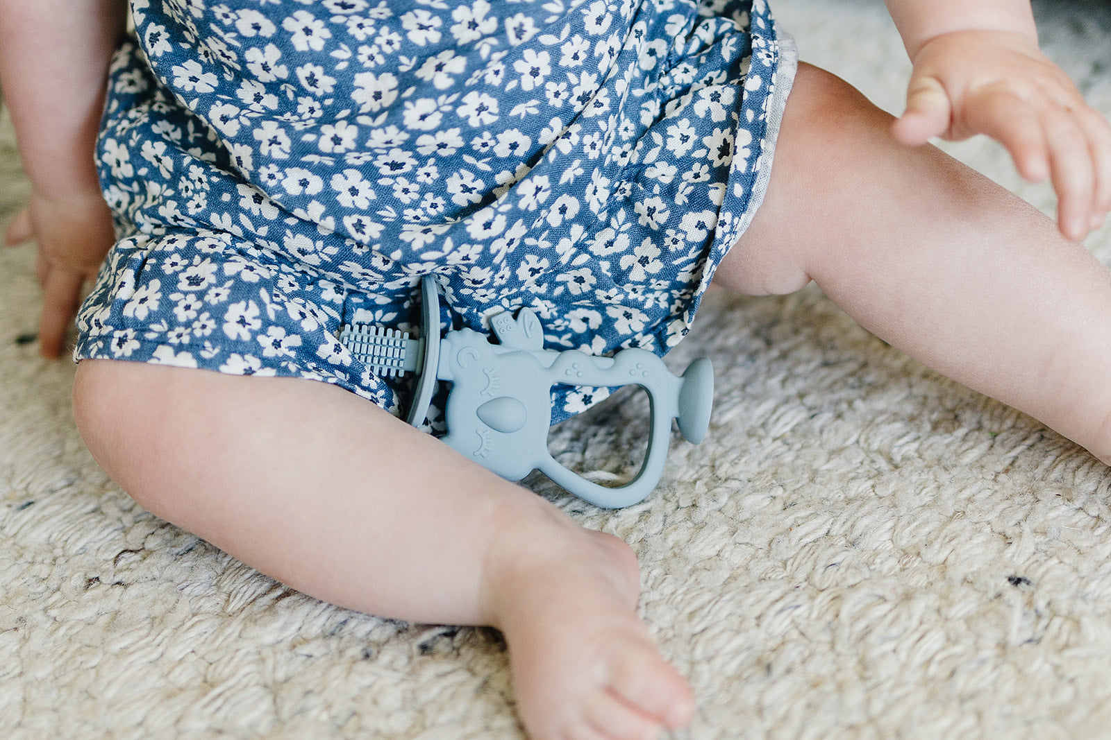 Baby wearing a blue floral outfit with a teething toy on a carpeted floor