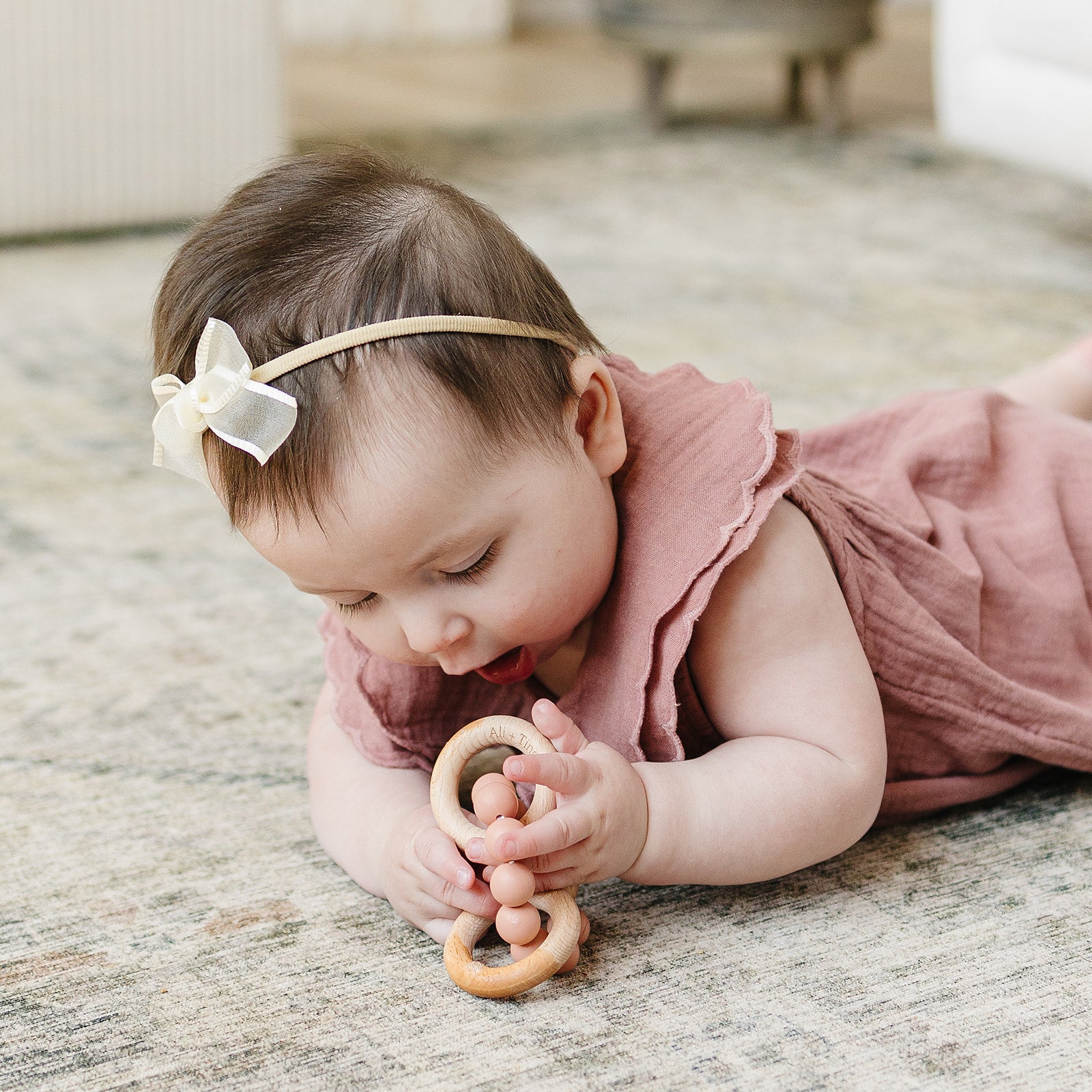 Baby lying on a wooden floor with a teething ring, wearing a pink dress and headband.