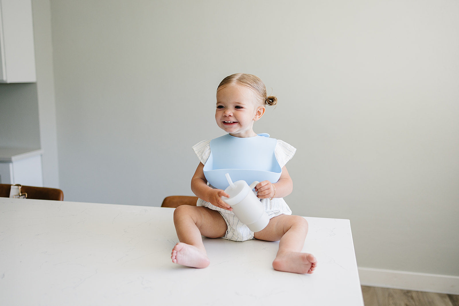 Child sitting on a table wearing a silicon bib and holding soft silicone tumbler for babies in neutral color in a room with white walls and furniture.