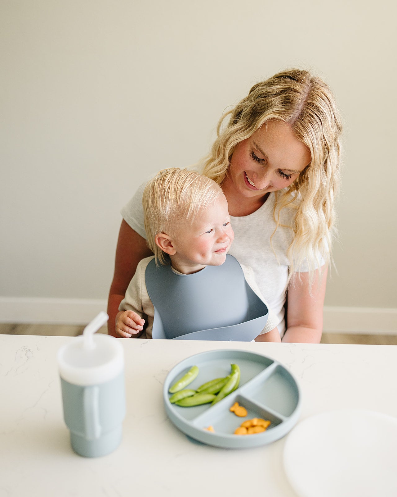 Woman with a child using a blue silicon bib, with a plate of food and a cup on a table.