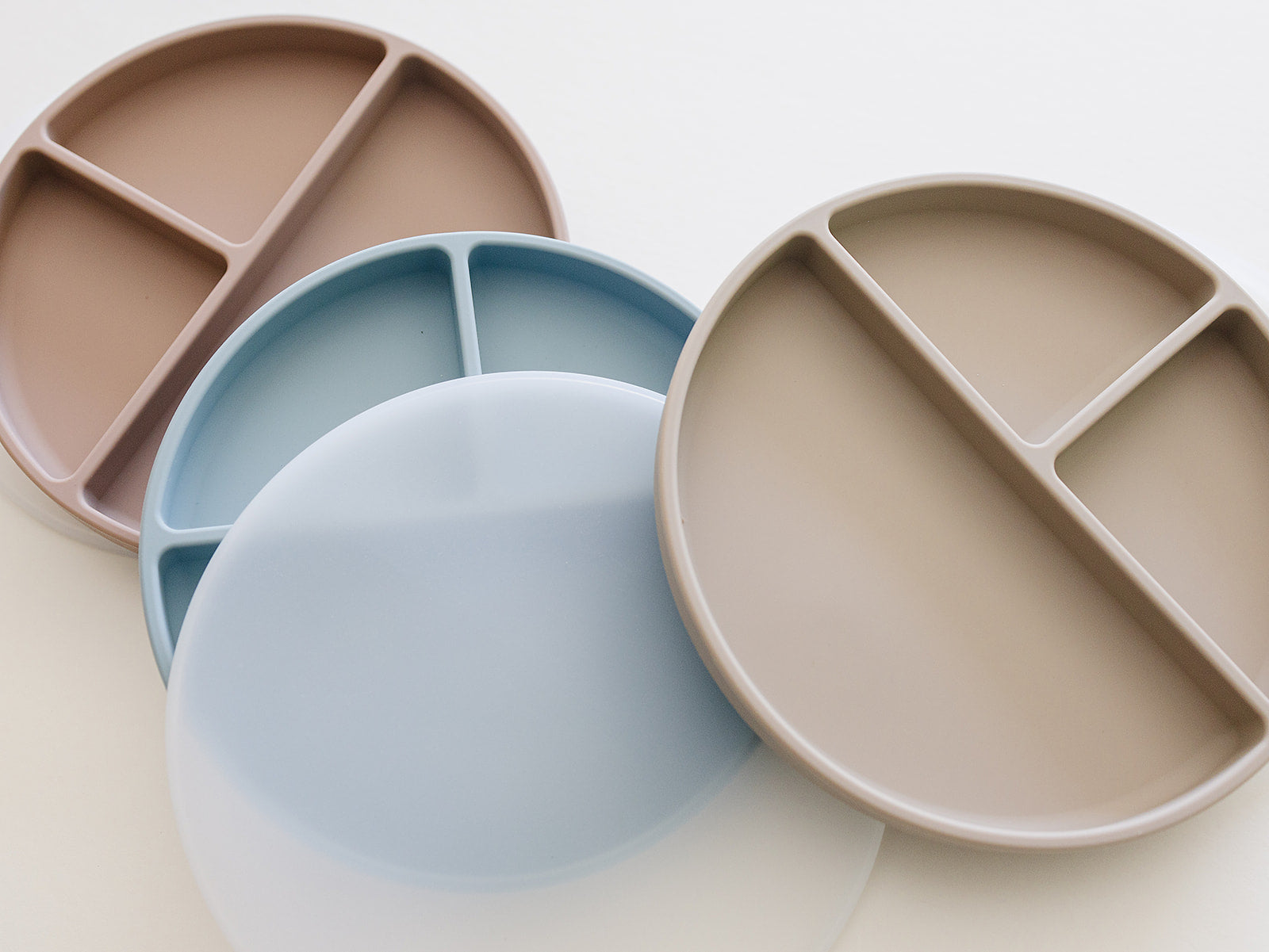 Three food-grade platinum silicone plates with food compartments in beige, blue, and pink on a light background