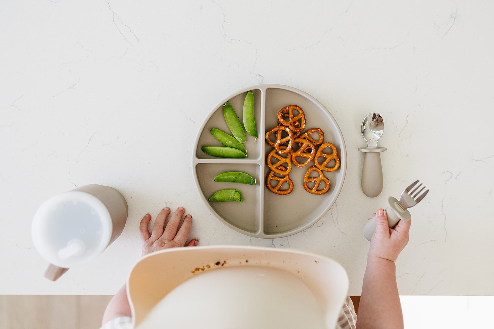 baby using divided silicone suction plate  with divided sections containing vegetables and pretzels, accompanied by a cup and cutlery on a white surface.