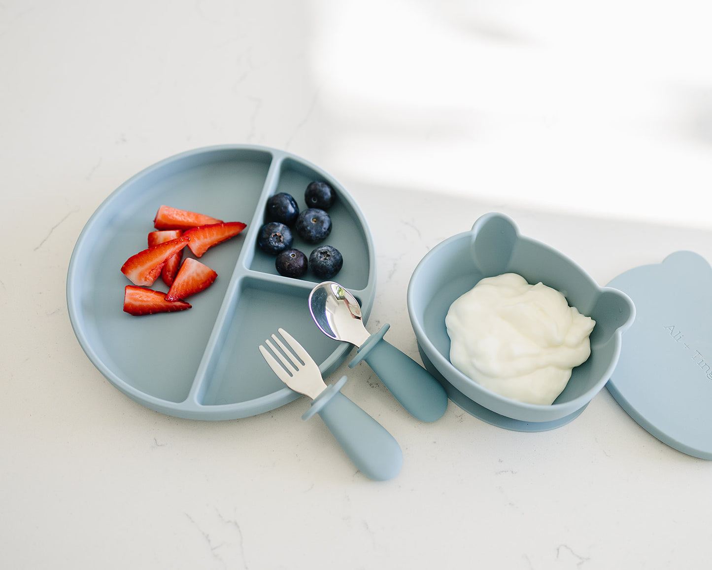 baby using divided silicone suction plate with divided sections, fork, and bowl with yogurt and fruit on a white background