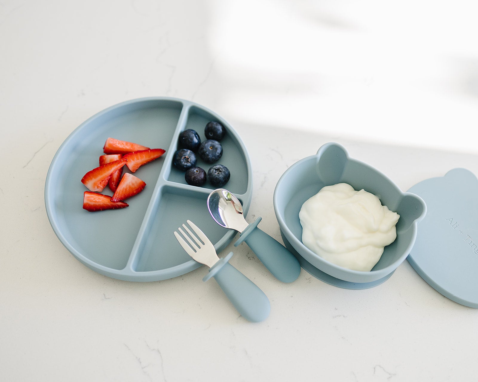 baby using divided silicone suction plate with divided sections, fork, and bowl with yogurt and fruit on a white background