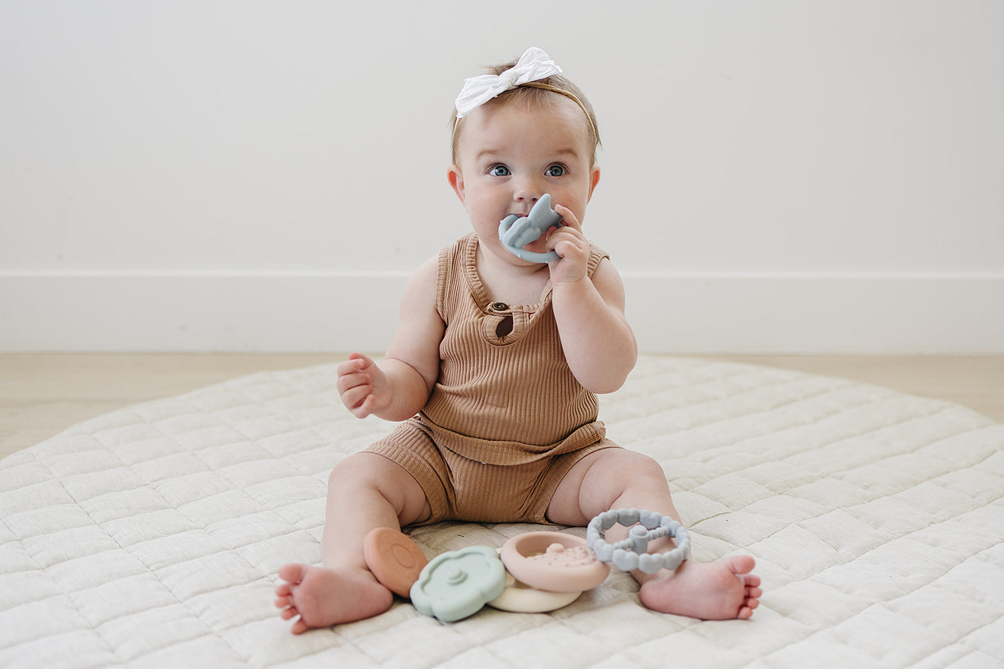 Baby sitting on a white blanket with colorful teething rings, wearing a brown onesie and white headband.
