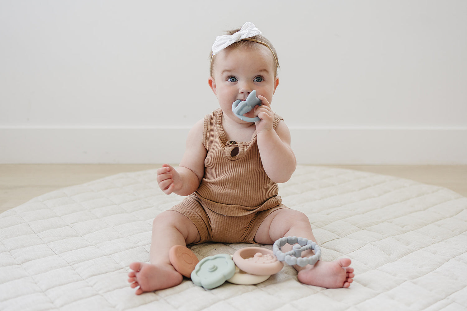 Baby sitting on a white blanket with colorful teething rings, wearing a brown onesie and white headband.