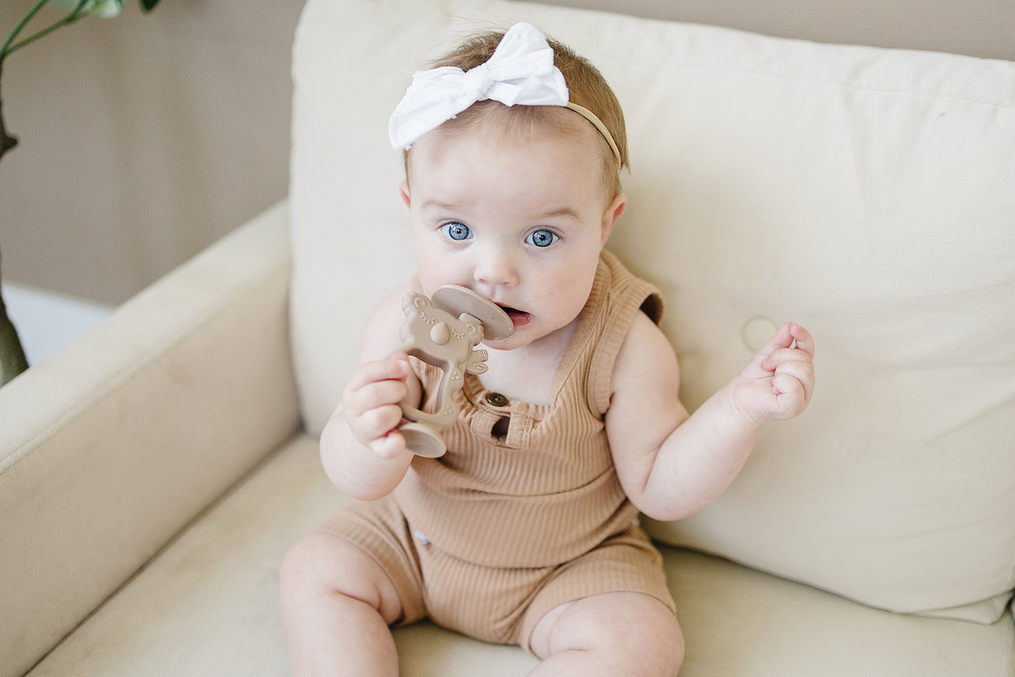 safe silicone teething toys Baby wearing a beige romper and white headband sitting on a light-colored couch.